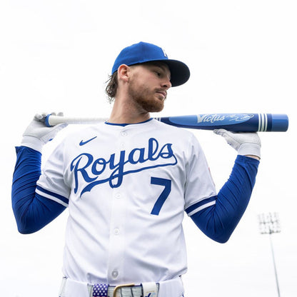 A baseball player in a white Kansas City Royals uniform and blue cap rests the 2025 Victus Vibe Pro-Crayon Bobby Witt Jr (-5) USSSA bat across his shoulders, gazing to the side with overcast skies and stadium lights behind him.