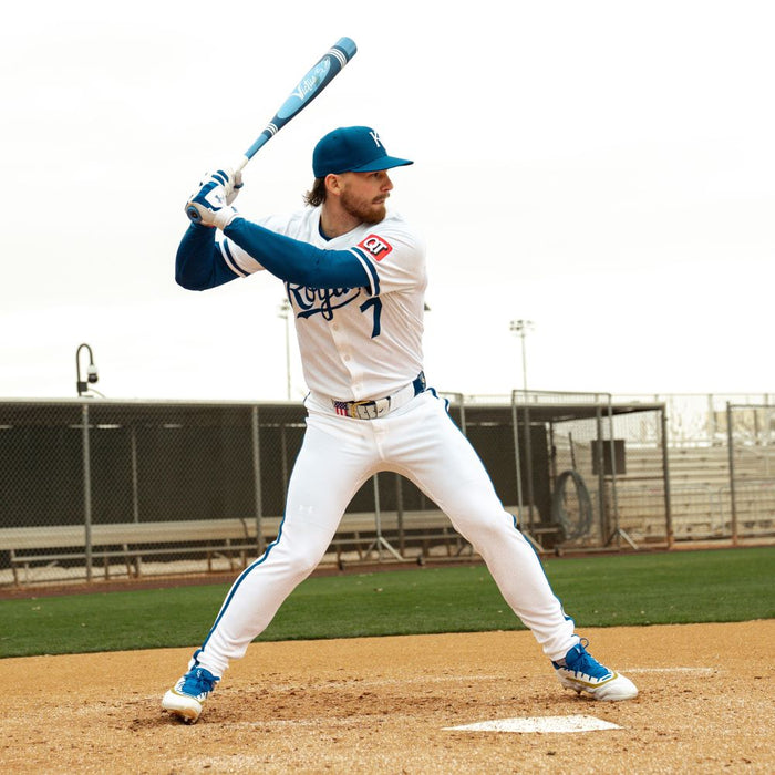 A baseball player in a white and blue uniform stands ready to bat at home plate, gripping the Victus 2025 Vibe Pro-Crayon Bobby Witt Jr (-5) USSSA Baseball Bat. A chain-link fence and field lights appear in the background.