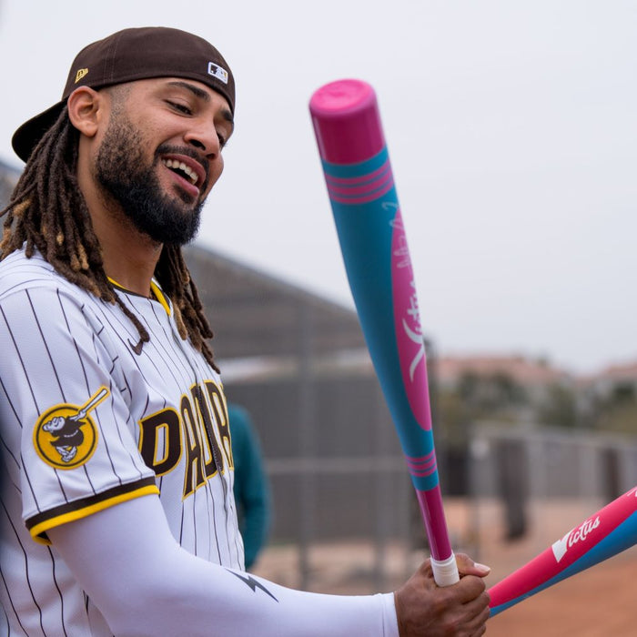 A Padres player in a backward cap smiles while holding the 2025 Victus Vibe Pro-Crayon Fernando Tatis Jr (-8) USSSA Baseball Bat, featuring a pink and blue design and vibration-reducing knob, during outdoor practice.