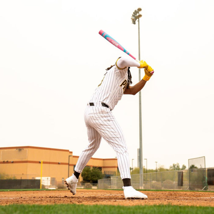A baseball player in a pinstripe uniform gets ready to bat on the field, gripping a Victus 2025 Vibe Pro-Crayon Fernando Tatis Jr (-8) USSSA bat with a vibration reducing knob and wearing yellow gloves; buildings and lights tower behind him.