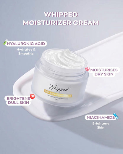 Whipped moisturizer cream in a clear jar with white cream visible, shown on a pale blue background with promotional text highlighting ingredients and skin benefits.