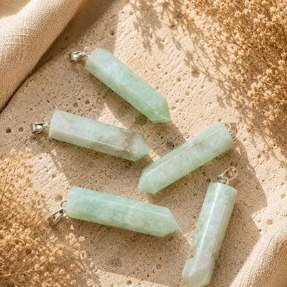 Five Amazonite point pendants with silver bail loops arranged on a textured beige stone surface with dried flowers and soft linen fabric.