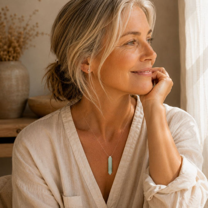 A woman with blonde hair in a low bun wears a delicate silver necklace with a light blue-green cylindrical pendant, resting her chin on her hand in a softly lit indoor setting.
