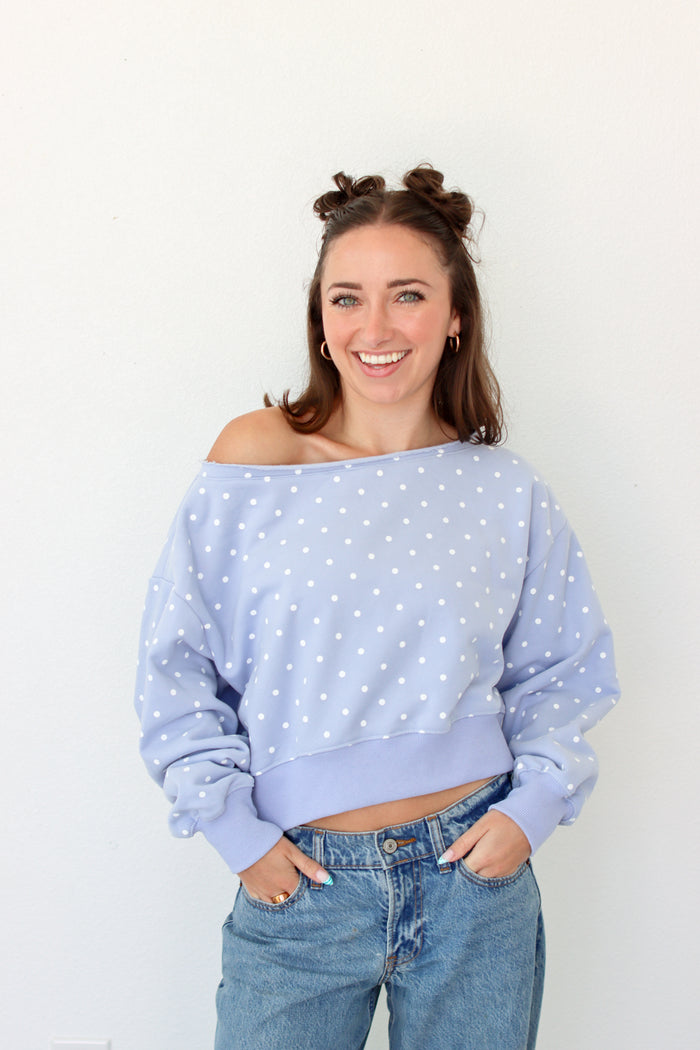 A smiling woman with her hair in two buns wears a light blue off-the-shoulder long-sleeve sweatshirt with white polka dots, paired with blue jeans, standing against a plain white wall.
