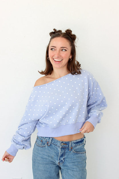 A woman wearing a light blue off-the-shoulder long-sleeve sweatshirt with white polka dots and blue jeans, standing against a plain white wall.
