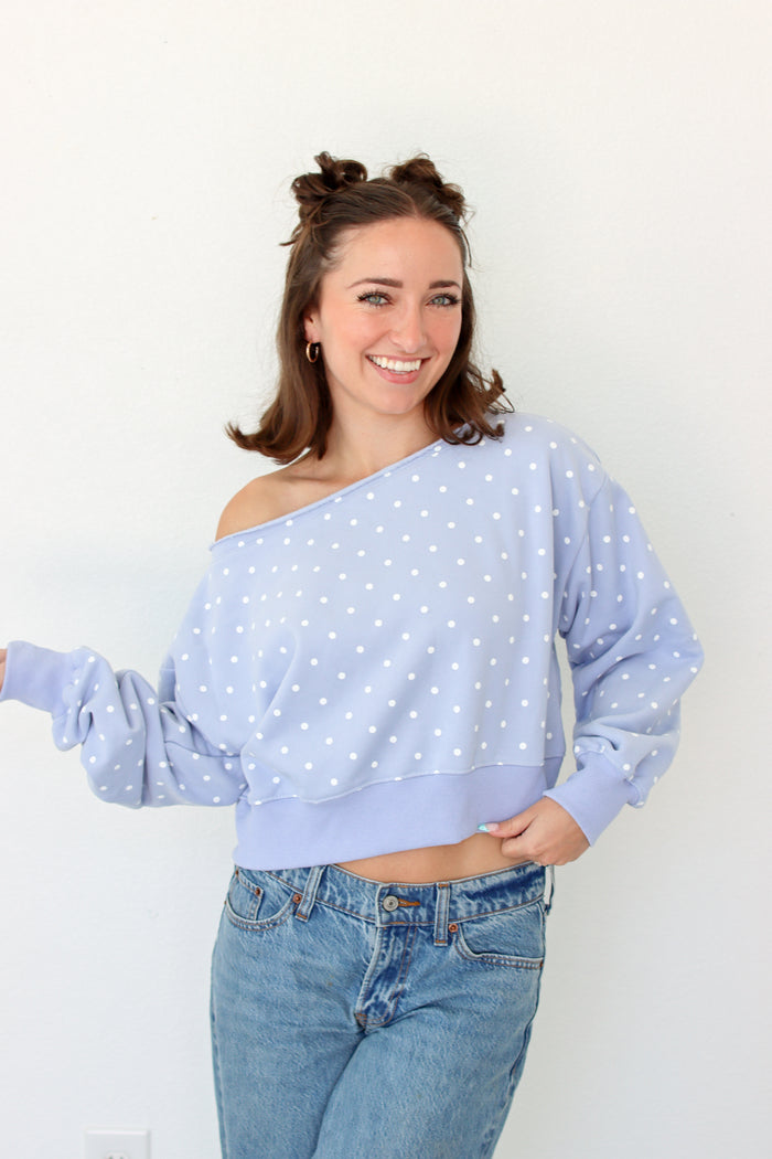 A woman modeling a light blue off-the-shoulder long-sleeve crop top with white polka dots, paired with blue jeans, against a plain white wall.