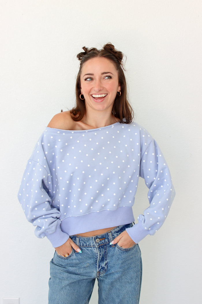 A woman wearing a light blue off-the-shoulder long-sleeve crop top with white polka dots and blue jeans, standing against a plain white wall.