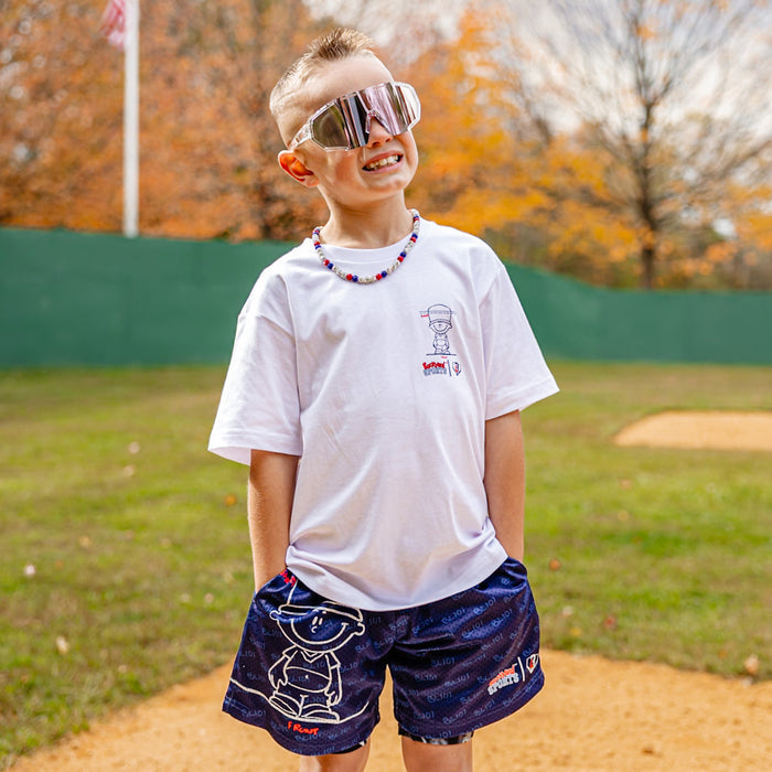 Child wearing sunglasses and a white Pablo Sanchez graphic t-shirt and blue Pablo Sanchez graphic shorts on a baseball field