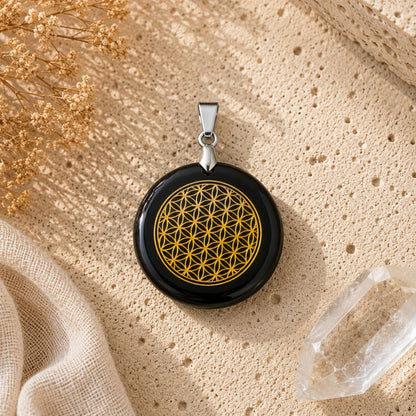 A round black pendant with a gold Flower of Life design, displayed on a textured beige fabric surface with dried flowers and a crystal.