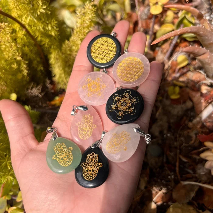 Hand holding a collection of crystal Metatron, Flower of Life, Hamsa, and Sacred Geometry pendants with gold symbols engraved on polished gemstone pendants.