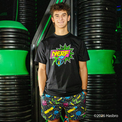 Person wearing a black t-shirt with a colorful 'NERF X BL101' logo and patterned shorts standing in front of green and background.
