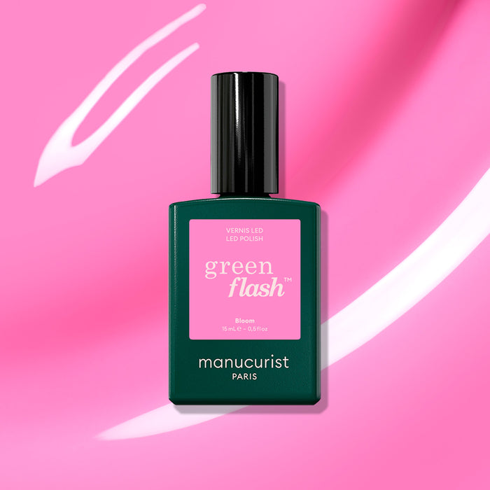 A bottle of Manucurist Bloom gel polish rests on a pink background with white streaks. The dark green bottle features a pink label marked “green flash” and “Bloom,” creating a magnetically intense look.