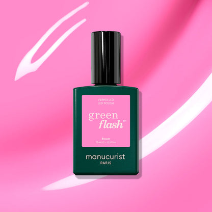 A bottle of Manucurist Bloom gel polish rests on a pink background with white streaks. The dark green bottle features a pink label marked “green flash” and “Bloom,” creating a magnetically intense look.