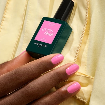 A hand with neatly manicured pink nails holds a bottle of Bloom gel polish by Manucurist, standing out against a yellow fabric background.