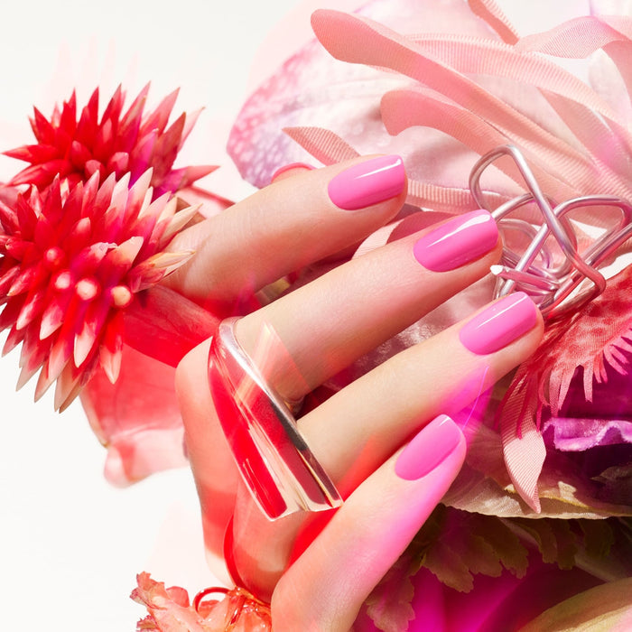 A vibrant close-up shows a hand with Manucurist's Bloom neon pink gel polish, styled with large silver rings and surrounded by vivid pink and red tropical flowers for a magnetic look.