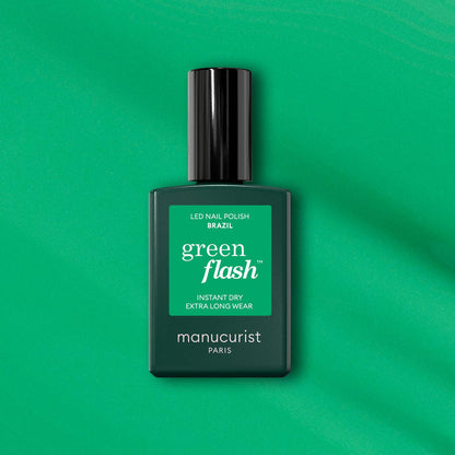 A bottle of Manucurist Brazil Green Flash LED polish sits on a green background, featuring instant dry and extra long wear for vibrant electric green nails inspired by Copacabana Beach.