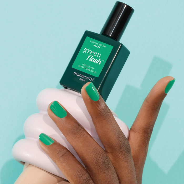 A hand with neatly manicured nails painted electric green holds a bottle of Manucurist Brazil polish against a light blue background.