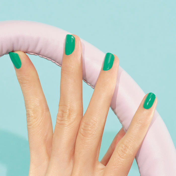 A hand with Manucurist's Brazil electric green nail polish holds a light pink, leather-textured steering wheel against a light blue background.