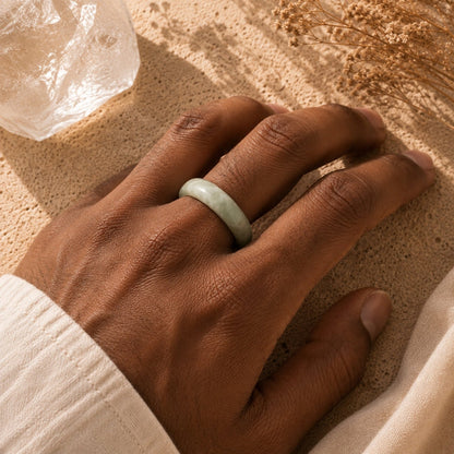 A light green jade bangle ring worn on a person's hand, resting on a textured sandy surface with dried flowers and a crystal cluster nearby.
