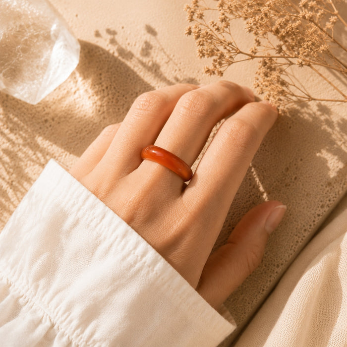 A close-up of a hand wearing a smooth, rounded Carnelian  ring, resting on a textured beige surface with dried flowers and warm sunlight.