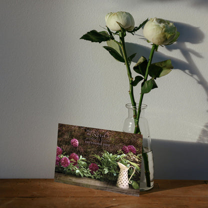 White roses in a glass vase next to a greeting card on a wooden surface with a light gray wall background.