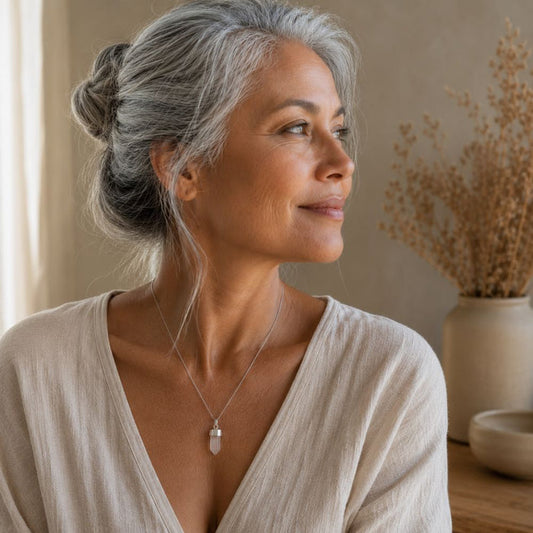 A close-up lifestyle shot of a woman with silver-gray hair in a bun, wearing a delicate silver pendant necklace with a clear crystal charm. She is wearing a beige linen top in a warm, softly lit room with dried flowers in the background.
