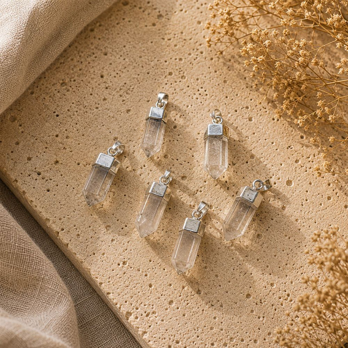 Five clear crystal point pendants with silver-toned caps and loops, arranged on a textured beige stone surface with linen and dried flowers in the background.