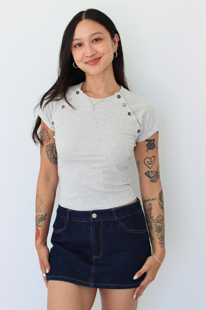 A woman modeling a light gray short-sleeve t-shirt with decorative buttons on the shoulders and a dark blue denim mini skirt, standing against a plain white wall.