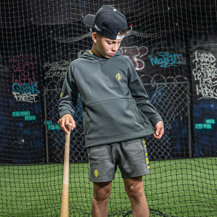 Child in gray hoodie and shorts with yellow BL101 logo, holding a baseball bat.