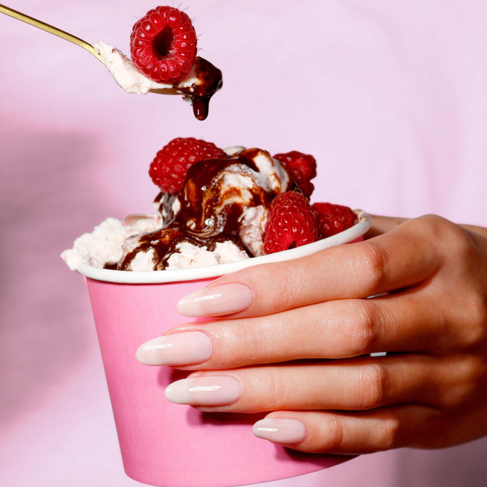 A hand with sheer manicured nails holds LONDONTOWN's Double Scoop—a pink cup of creamy ice cream topped with raspberries and chocolate sauce—as a fork lifts a raspberry.