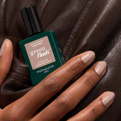 A hand with neatly manicured pastel beige nails holds a dark green Manucurist bottle of Dove Beige gel polish, resting on brown leather.