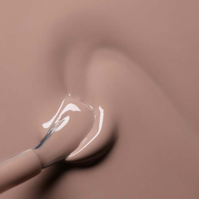 A close-up of a Manucurist nail polish brush applying glossy Dove Beige gel polish in smooth strokes on a matching background.