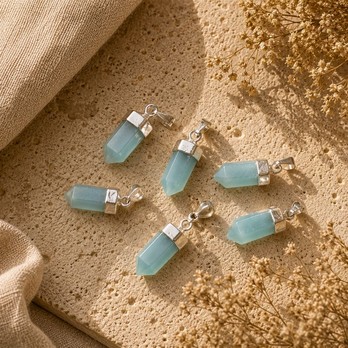 Five light blue gemstone pendant charms with silver-toned caps and lobster clasps, arranged on a textured beige stone surface with burlap and dried flowers.
