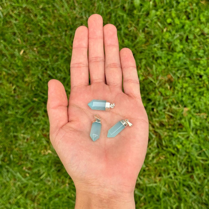 Three small light blue crystal point pendants with silver jump rings are held in the palm of a hand, set against a blurred green grass background.
