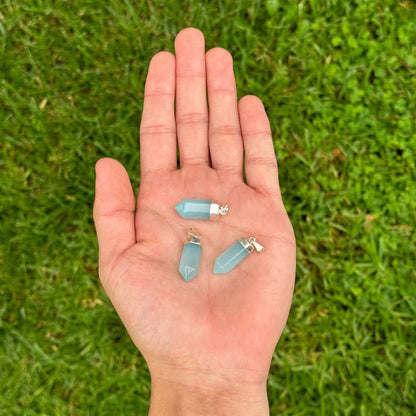 Three small light blue crystal point pendants with silver jump rings are held in the palm of a hand, set against a blurred green grass background.