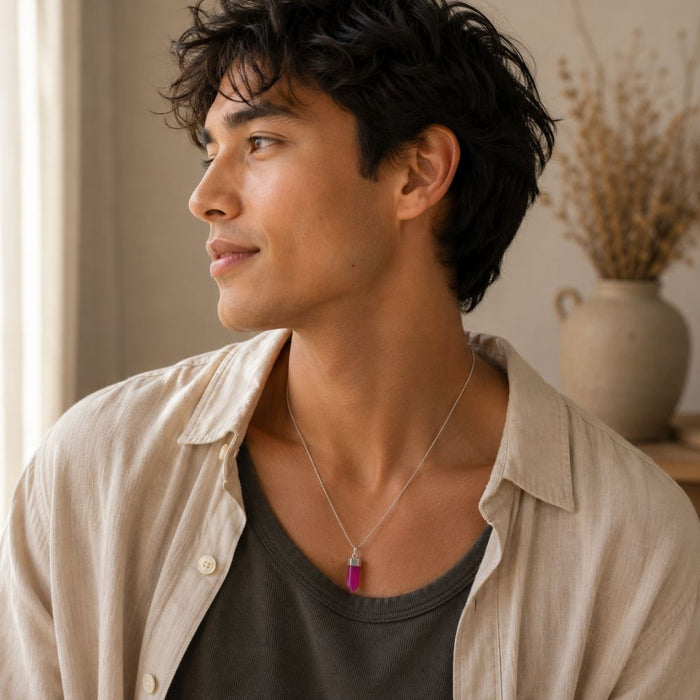 A man wearing a silver-toned pendant necklace with a vibrant magenta-pink rectangular stone, styled with a beige linen shirt over a dark gray t-shirt in a softly lit indoor setting.