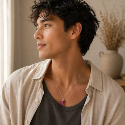 A man wearing a silver-toned pendant necklace with a vibrant magenta-pink rectangular stone, styled with a beige linen shirt over a dark gray t-shirt in a softly lit indoor setting.