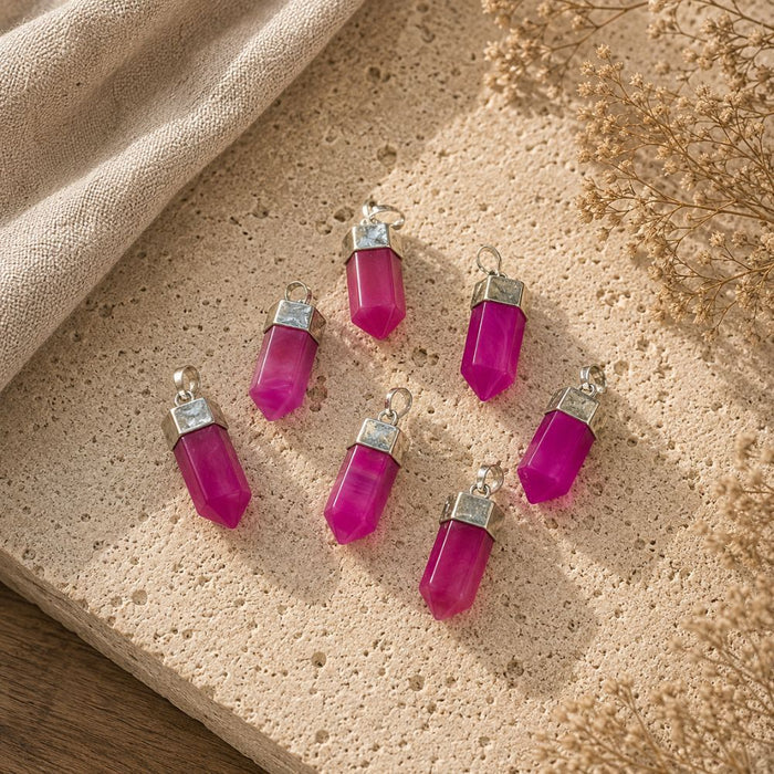 Five magenta-pink crystal pendants with silver caps and loops, arranged on a textured beige stone slab with a linen cloth and dried flowers.