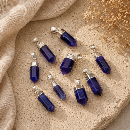 Six blue crystal point pendants with silver caps and loops, arranged on a textured beige stone surface with a beige fabric drape and dried flowers.