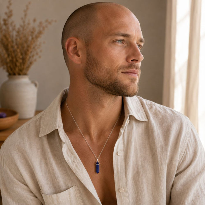 A man wearing a silver-toned pendant necklace with a dark blue rectangular stone, styled in a warm, sunlit indoor setting.