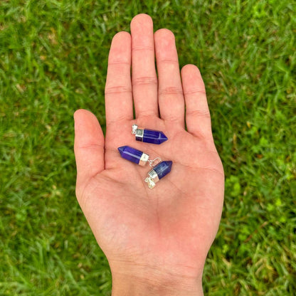 Three small cobalt blue crystal point pendants with silver caps are displayed on an open hand, set against a blurred green grass background.