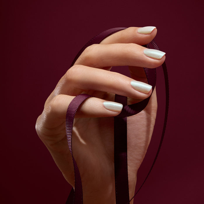 A hand with neatly manicured Fairy nail polish by Manucurist holds a dark purple ribbon against a matching burgundy background.