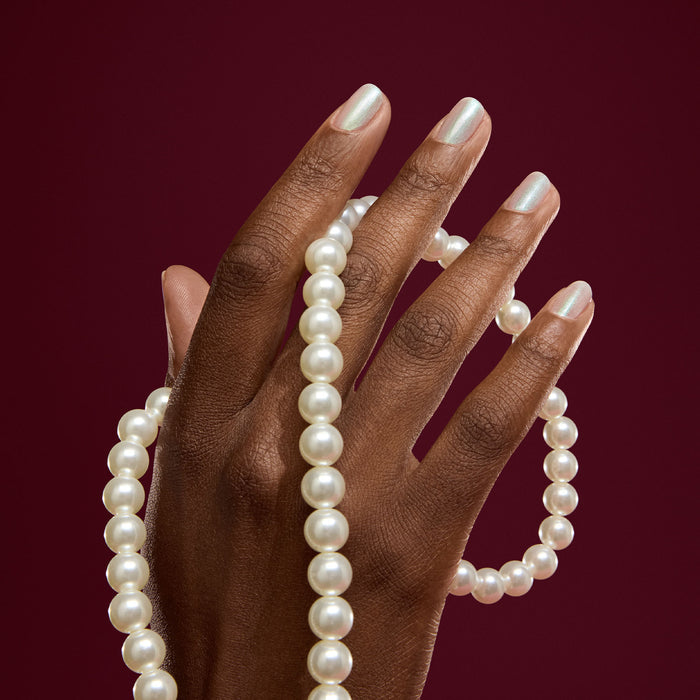 A hand with Manucurist’s Fairy pearly white nail polish holds a string of white pearls against a dark maroon background.