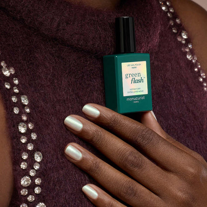 A hand with pearly white gel polish rests on a purple textured top with shiny rhinestones, holding a teal bottle of Fairy nail polish by Manucurist for ultimate sparkle nails.
