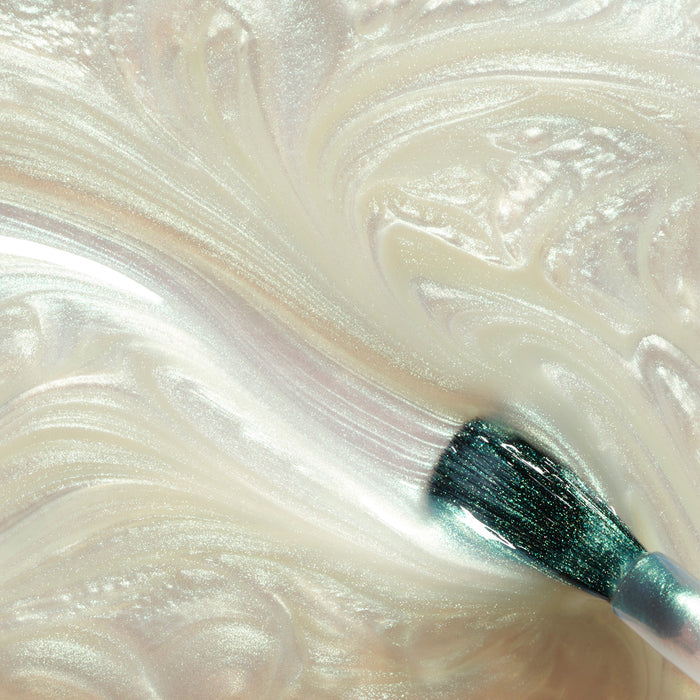 A close-up of a makeup brush swirling through Manucurist's Fairy, a shimmery, pearlescent cream, creating smooth iridescent waves—evoking the look of pearly white nail polish.