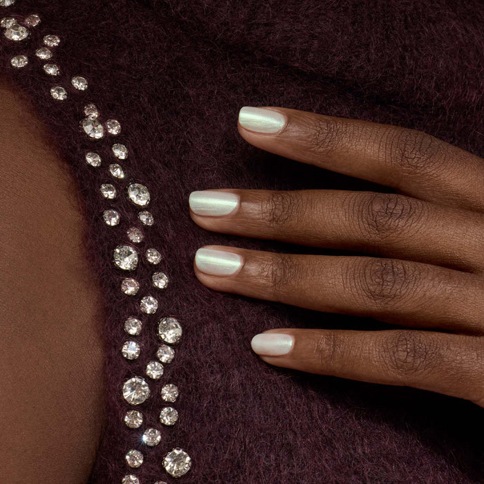 A hand with Manucurist's Fairy pearly white nail polish rests on dark purple textured fabric, adorned with rows of sparkling round rhinestones.
