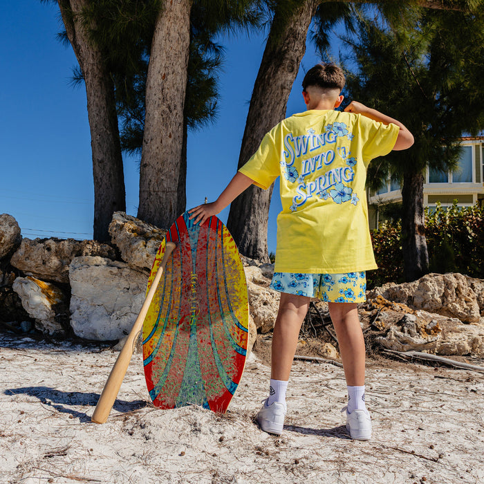 Person in yellow shirt with 'Swing into Spring' text and floral shorts holding a colorful board on a beach.