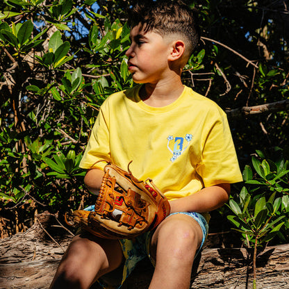 Child in a yellow floral BL101 shirt holding a baseball glove in a natural setting