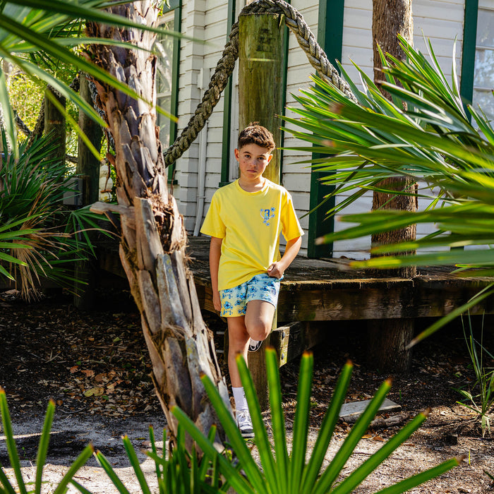 Person wearing a yellow BL101 shirt and blue floral shorts standing in a tropical setting with plants and a house in the background.