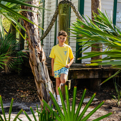 Person wearing a yellow BL101 shirt and blue floral shorts standing in a tropical setting with plants and a house in the background.